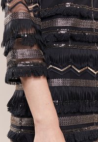 Robe noire avec des détails frangés, des manches transparentes et des rangées alternées de sequins et d'accents métalliques, présentant des motifs texturés.