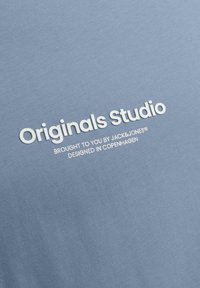 Camisa de algodón azul con una textura suave que presenta texto blanco que dice "Originals Studio" y detalles adicionales de marca debajo.