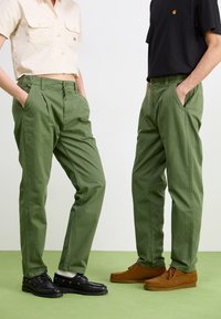 Green cotton trousers with pleats, side pockets, and contrasting brown suede loafers. Black lace-up shoes complement the outfit.