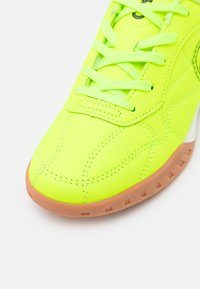 Neon geltonos sportbačiai su tekstūruotu sintetinės medžiagos viršumi, toninėmis siūlėmis, raišteliais ir šviesiai rudos spalvos padu su mažais sukibimo raštais.