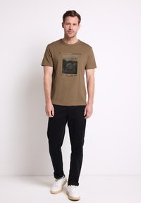 Man i brun grafisk t-shirt, svarta byxor och vita sneakers som står mot en enkel vit bakgrund.