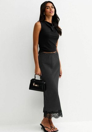 Vrouw draagt een mouwloze zwarte top en een zwarte rok met witte stippen en een kanten zoom, houdt een zwarte handtas vast en draagt zwarte sandalen met hak.
