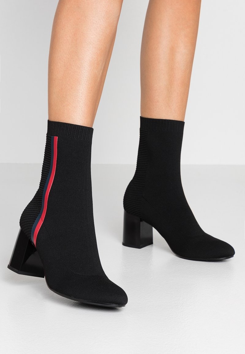 Botas de tobillo de punto negro con punta afilada, textura acanalada y tacón cuadrado. Presentan una franja lateral en rojo y azul.