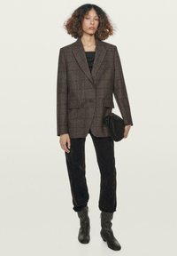 Blazer a quadri marrone con rever a punta, chiusura con un bottone, abbinato a pantaloni scuri slim-fit e stivaletti neri con tacco. Tiene una clutch nera.