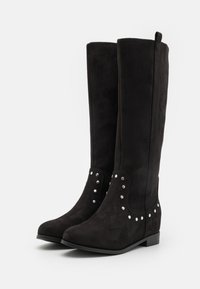 Botas negras hasta la rodilla, hechas de ante, con suela plana y pequeños clavos plateados redondos a lo largo del costado para aportar detalle.