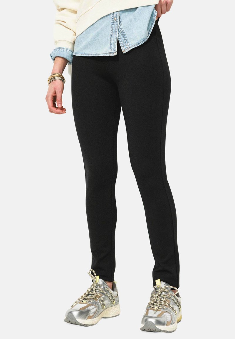 Leggings noirs en tissu extensible avec une coupe slim, présentant une texture lisse et des coutures latérales contrastées, associés à des sneakers argentées.