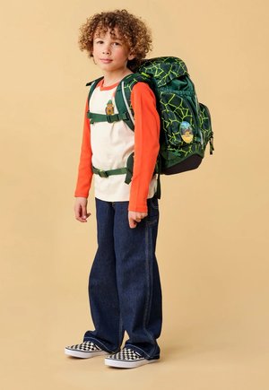 Kind mit lockigem Haar, trägt ein oranges und weißes Hemd, blaue Jeans, karierte Slip-On-Schuhe und einen grünen Rucksack mit Muster und Dino-Anstecker.
