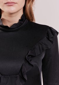 Černá blůza s vysokým límcem a volánkovým detailem podél ramenního švu. Vyrobená z hladké, strukturované látky.