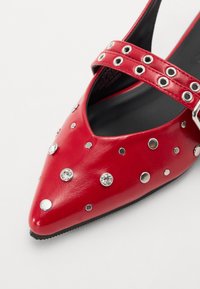Chaussure à talons hauts en cuir rouge avec bout pointu, ornée de clous en argent et de strass, ainsi qu'une large sangle avec découpes circulaires.