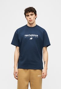 Νεαρός άνδρας που φοράει μπλε σκούρο μπλουζάκι New Balance Athletics και καφέ μπεζ παντελόνι φόρμας New Balance, στέκεται μπροστά σε ένα απλό φωτεινό φόντο.