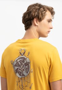 T-shirt in cotone giallo con una grafica di un orologio da tasca sul retro, caratterizzato da dettagli intricati e con i numeri 12, 3, 6 e 9 ben visibili.