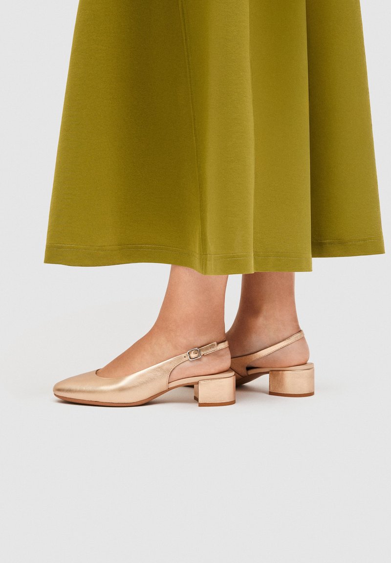 Ben iført guldfarvede slingback-sko med blokhæl, kombineret med en grøn midinederdel, vist mod en ensartet lys baggrund.