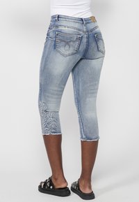 Personne portant un jean bleu clair court avec un motif floral brodé sur une jambe et des sandales noires à enfiler avec des clous argentés.
