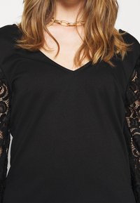 Musta paita, jossa on V-pääntie ja pitsiset pitkät hihat, sileä tekstuuri ja monimutkainen pitsikuvio hihojen reunoilla. Mukana kultaketjun kaulakoru.