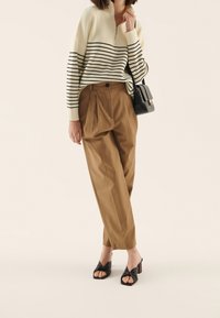 Suéter crema con rayas negras, cuello con cremallera; pantalones marrones de pierna ancha con pliegues; tacones abiertos negros con nudos; bolso pequeño negro.