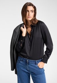 Schwarzes Satin-Hemd mit Knöpfen und langen Ärmeln, kombiniert mit ausgebleichten blauen Jeans. Grauer Blazer über die Schulter drapiert.