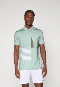Polo in tessuto verde chiaro, caratterizzato da un design geometrico con linee diagonali e righe in verde scuro e bianco. Maniche corte.