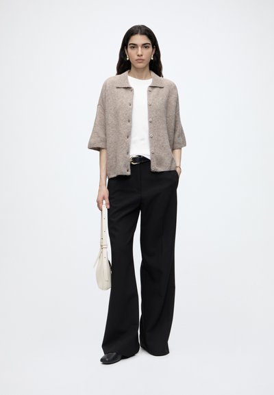 Femme portant un cardigan beige à manches courtes boutonné sur une chemise blanche, pantalon noir à jambes larges, chaussures noires, tenant un sac à main blanc sur fond uni.