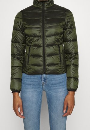 Vrouw draagt een nauwsluitend olijfgroen doorgestikt jack met ritssluitingen op de zakken en een blauwe spijkerbroek, staand tegen een effen achtergrond.