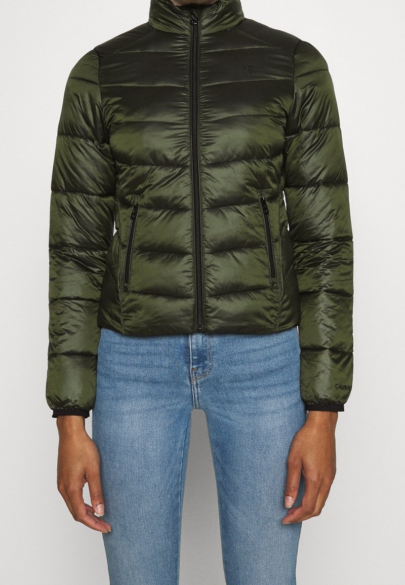 Femme portant une veste matelassée ajustée de couleur vert olive avec des poches zippées et un jean bleu, debout devant un fond uni.