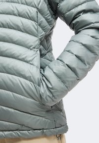 Doudoune matelassée gris clair avec des coutures horizontales, portée les mains dans les poches latérales, pantalon beige visible sous l’ourlet de la veste.