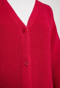 Cardigan rouge en tricot avec un col en V, doté de deux boutons ronds et d'un tissu texturé. Vue en gros plan mettant en valeur le motif en maille.