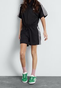 Person wearing black Adidas short-sleeve jumpsuit with white stripes and green Adidas sneakers with pink socks, standing against plain wall.