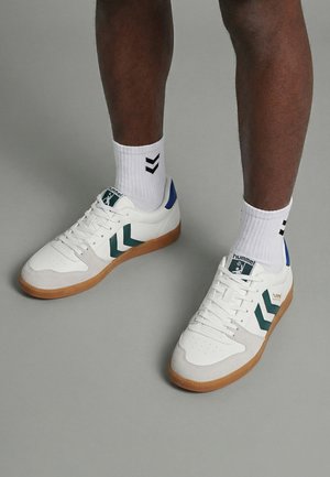 Nogi w białych butach z zielonymi akcentami w kształcie zygzaka i gumowymi podeszwami, uzupełnione białymi skarpetkami z dwoma czarnymi zygzakami po obu stronach.