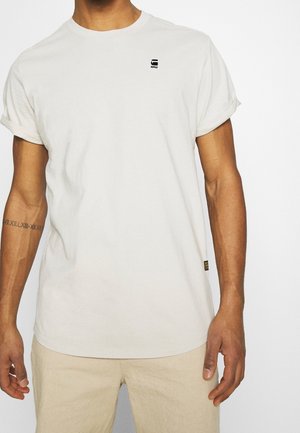 Homme portant un t-shirt uni blanc cassé avec un petit logo noir sur la partie supérieure gauche de la poitrine et un pantalon beige, tatouage visible sur l'avant-bras gauche.