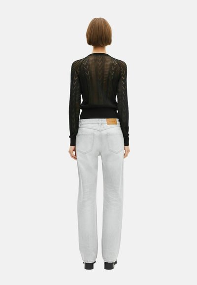 Mujer con cabello corto castaño, vistiendo una blusa negra transparente de manga larga con un patrón y jeans rectos de lavado claro, de espaldas al espectador.