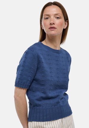 Femme portant un pull tricoté bleu à manches courtes avec un motif texturé et un pantalon écru à fines rayures verticales, sur un fond uni.