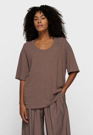 Kobieta o kręconych włosach, ubrana w luźną brązową koszulę z krótkim rękawem i pasujące szerokie spodnie, stojąca z jedną ręką w kieszeni.
