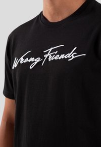Camiseta de algodón negra con texto bordado en blanco "Amigos Equivocados". Mangas cortas, cuello redondo clásico, textura suave.