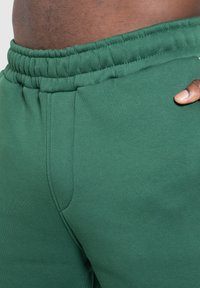 Nærbillede af en mørkhudet person iført bukser med grøn elastisk talje, med hånden hvilende nær hoften mod en lys baggrund.