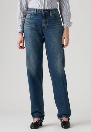 Femme portant un jean bleu droit, des chaussures noires à talons bas et une chemise rayée à manches longues avec manches retroussées.