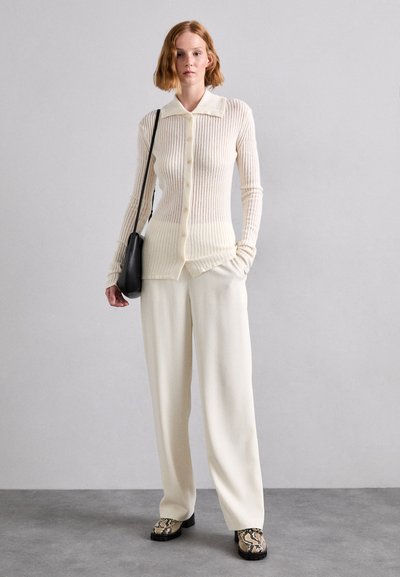 Cardigan crem cu dungi, cu guler și nasturi, asociat cu pantaloni crema cu pantaloni largi și pantofi negri cu un model de șarpe.