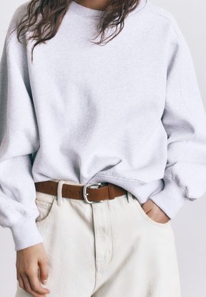 Persona que lleva una sudadera gris claro metida en unos pantalones blanco roto con un cinturón de cuero marrón, una mano en el bolsillo, cabello oscuro suelto visible.