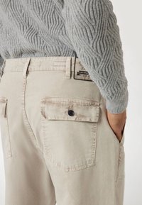 Beige utility pants met knoopzakken op de rug, een gestructureerde stof en een zichtbare merklabel op de tailleband. Draag ze met een lichtgrijze gebreide trui.