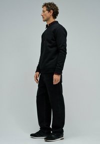 Sudadera negra con texto en relieve, combinada con jeans anchos negros. El modelo lleva zapatillas negras con toques marrones.