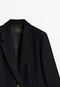 Manteau noir sur mesure avec larges revers, un seul bouton visible et doublure intérieure noire sur fond blanc.