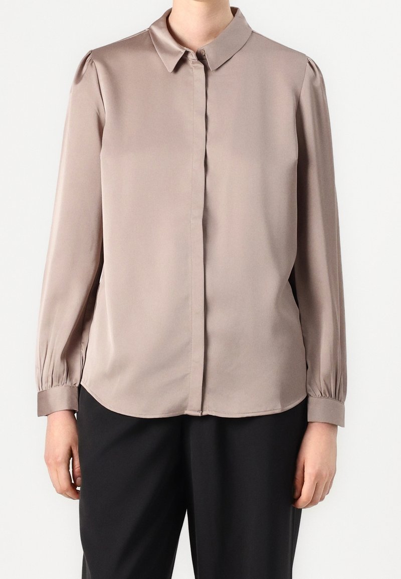 Personne portant un chemisier taupe à manches longues avec un col pointu et des boutons dissimulés à l'avant, associé à un pantalon noir.