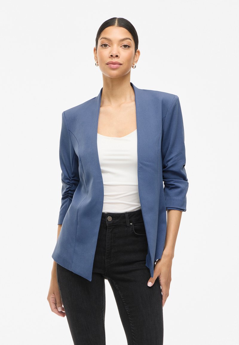 Blå blazer med en glatt tekstur, åpen front og 3/4-ermer, kombinert med en hvit topp og svarte jeans.