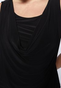 Svart ärmlös topp med draperad tyg i en lager-på-lager, cowl neck-design. Slät yta och mjuka veck skapar ett flytande utseende.