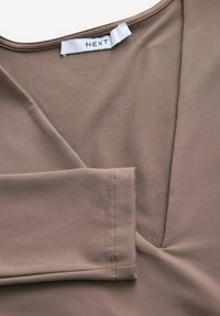 Bruine v-hals top gemaakt van een gladde stof, met stikdetails en een label met de tekst "NEXT." De stofval toont een zachte textuur.