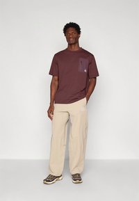 Bordeauxrood T-shirt met een borstzakje aan de linkerkant, beige broek en donkere sneakers met een lichte zool. Eenvoudig ontwerp, korte mouwen en een ontspannen pasvorm.