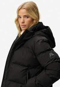 Femme blonde portant une veste matelassée noire de Superdry avec capuche, se retournant sur son épaule devant un fond blanc.