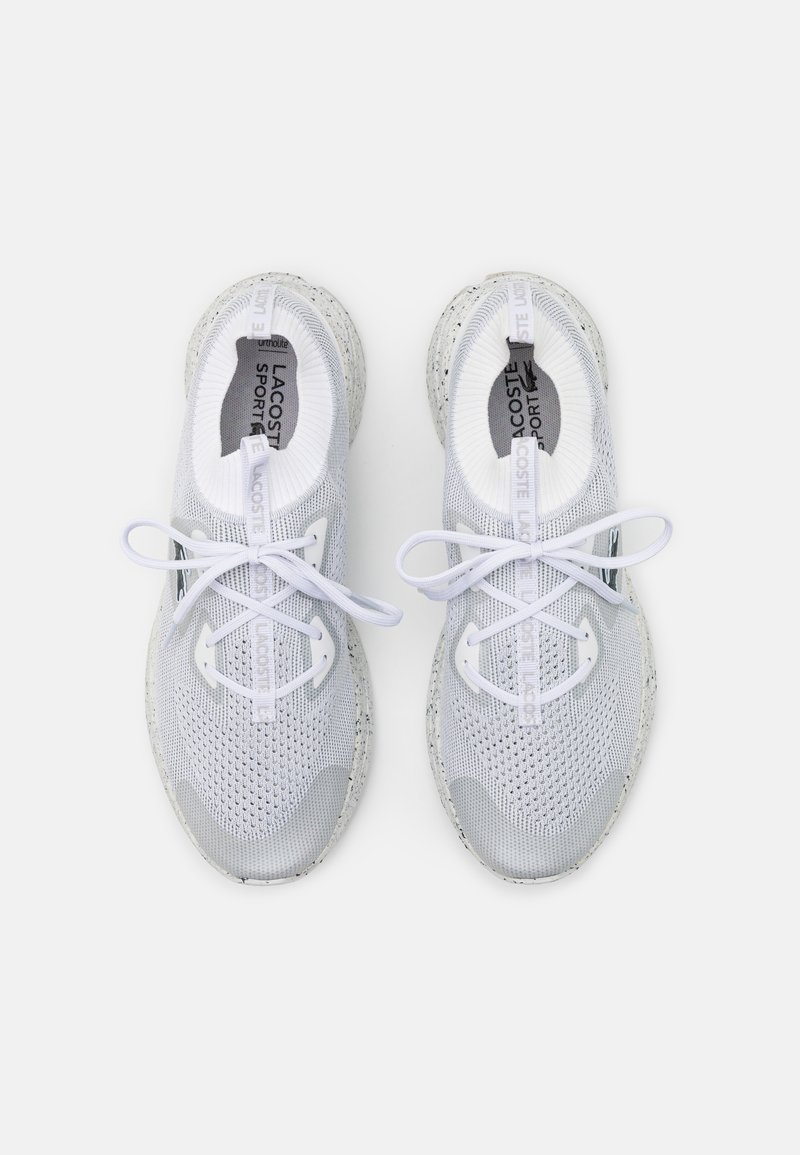 Une paire de baskets Lacoste Sport blanches avec une texture tricotée, un design à lacets et une semelle mouchetée, vue de dessus sur un fond blanc.