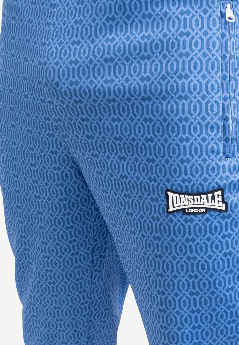 Blaue Sporthose mit geometrischem Muster, einer Reißverschlusstasche und einem auffälligen Lonsdale-Logo auf der linken Seite. Glatte Textur.