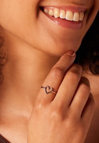 Zilveren ring met een hartontwerp, gedragen om een vinger. De band is dun en glad, wat een minimalistische stijl benadrukt. Huidtint zichtbaar.
