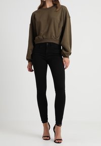 Mujer vestida con sudadera de manga larga verde oliva, jeans ajustados negros y tacones de tiras en el tobillo de punta abierta, de pie contra un fondo liso.